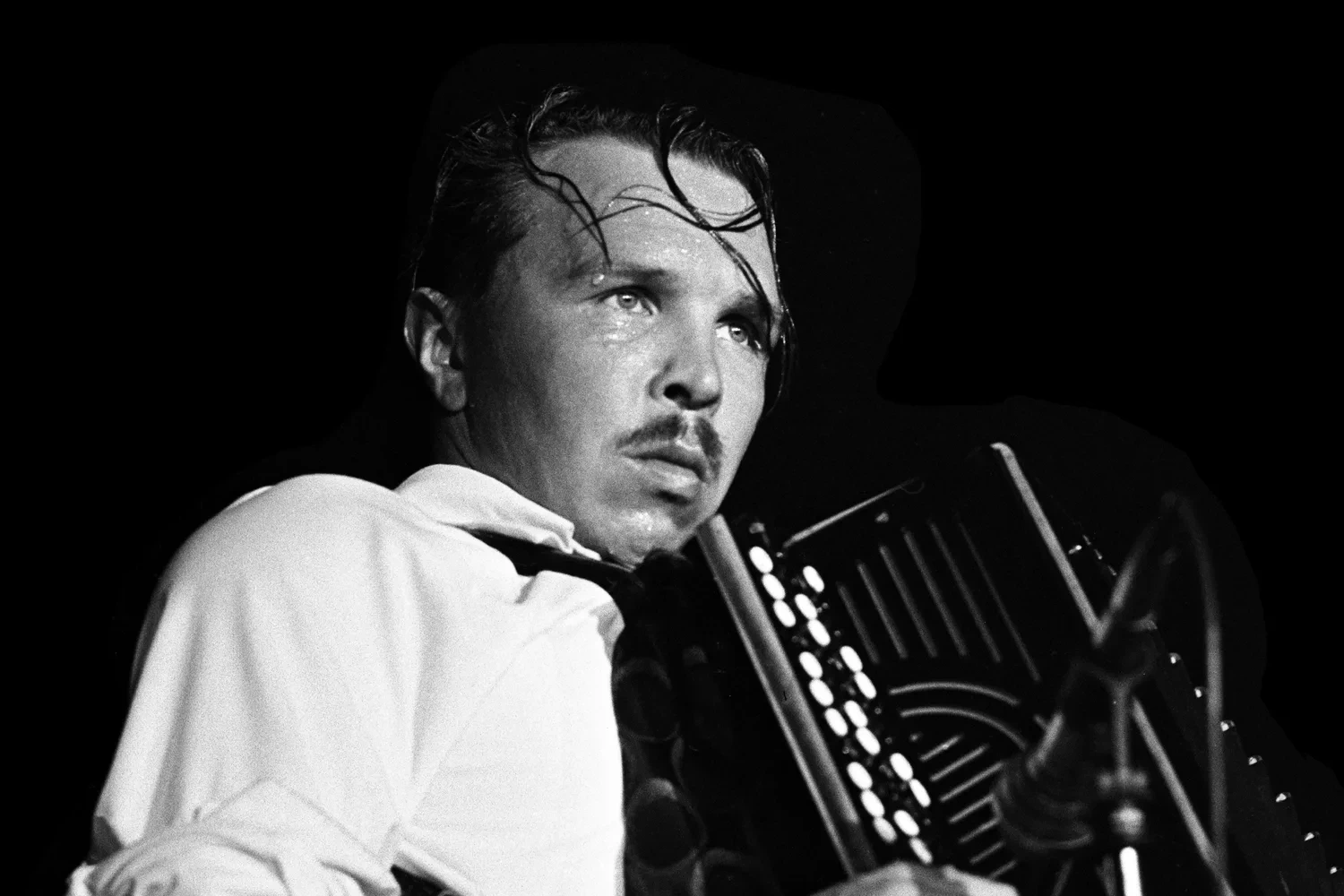 Black-and-white stage photo of Garik Sukachev, sweat on his face, playing a button accordion beside a microphone stand.