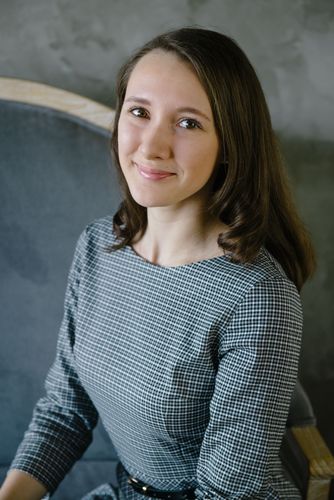 Smiling young woman with long brown hair wearing a grey houndstooth dress, seated on an armchair.