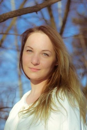 Blonde woman smiling outdoors with bare tree branches in the background.