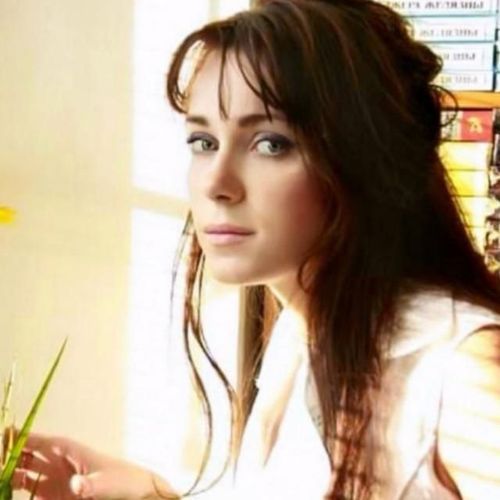 Young woman with long dark hair and bangs in a white top looking towards the camera next to a bookshelf.