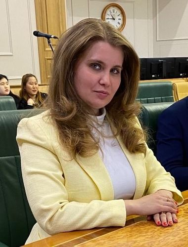 Woman in a yellow blazer seated at a conference table in an assembly room, with other participants behind her.