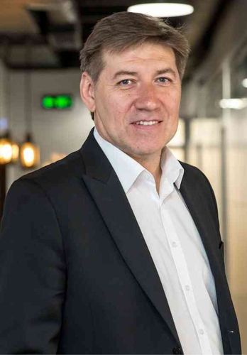 Smiling man with short brown hair in a black blazer and open-collar white shirt