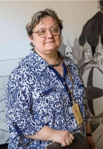 Woman with glasses in a blue patterned blouse standing before an illustrated wall